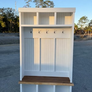 May include: White wooden entryway bench with a built-in shelf and hooks. The bench has a dark wood seat and three cubby storage spaces below. The top shelf has three open compartments. The hooks are black.