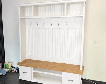 May include: A white wooden entryway bench with a natural wood seat and a built-in shelf. The bench has two drawers and six hooks for hanging items. Above the hooks are three small shelves.