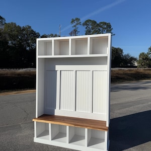 May include: A white wooden entryway bench with a natural wood seat and storage cubbies below. The bench has a vertical panel back and open shelves above. The bench is designed for organization and storage.