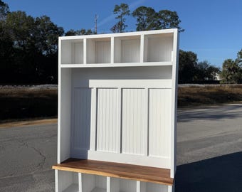 May include: A white wooden entryway bench with a natural wood seat and storage cubbies below. The bench has a vertical panel back and open shelves above. The bench is designed for organization and storage.