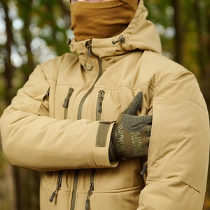 May include: A tan-colored jacket with a hood and a zipper closure. The jacket has multiple pockets and a velcro patch on the sleeve. The jacket is worn by a person wearing a brown balaclava.