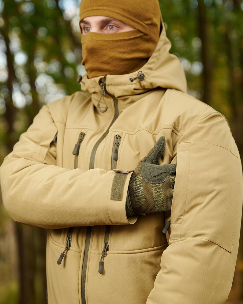 May include: A tan-colored jacket with a hood and a zipper closure. The jacket has multiple pockets and a velcro patch on the sleeve. The jacket is worn by a person wearing a brown balaclava.
