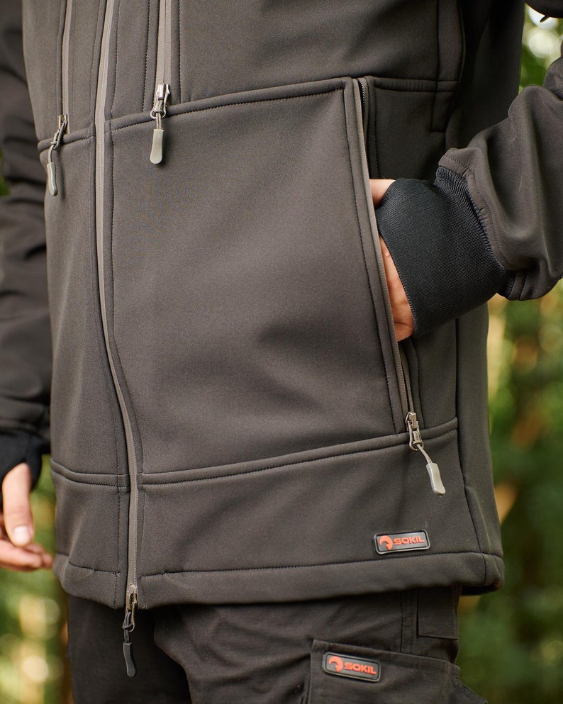 May include: A dark gray softshell jacket with a zipper closure and multiple pockets. The jacket has a logo on the bottom right pocket that says "SOKIL".
