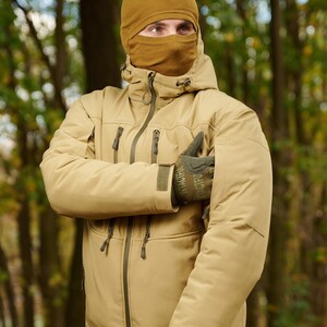 May include: A tan-colored jacket with a zipper and multiple pockets. The jacket has a hood and is worn by a person wearing a brown balaclava.
