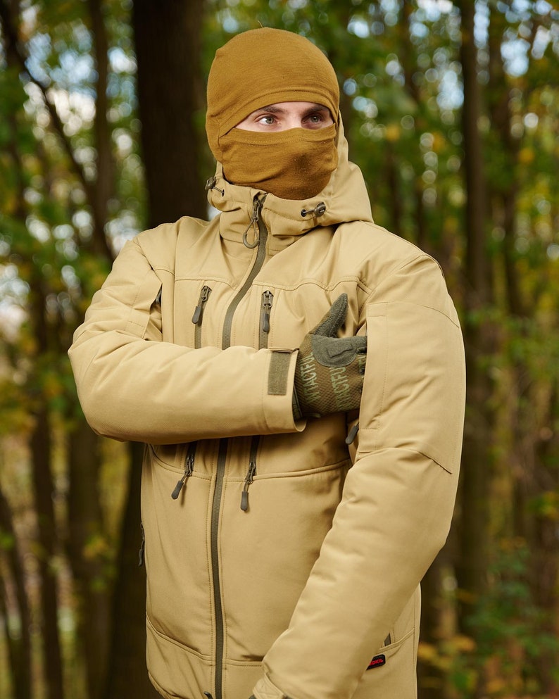 May include: A tan-colored jacket with a zipper and multiple pockets. The jacket has a hood and is worn by a person wearing a brown balaclava.