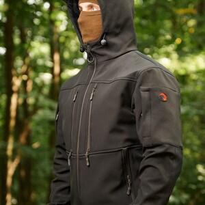 May include: A black softshell jacket with a zipper closure and multiple pockets. The jacket has a hood with a drawstring and a logo on the sleeve.