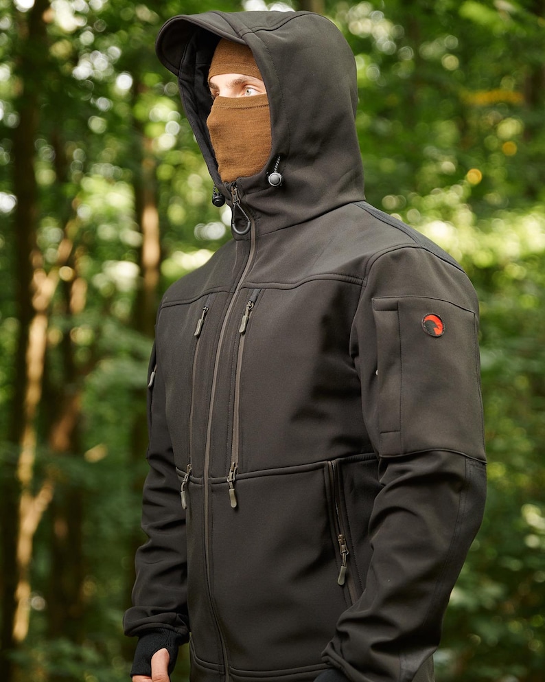 May include: A black softshell jacket with a zipper closure and multiple pockets. The jacket has a hood with a drawstring and a logo on the sleeve.