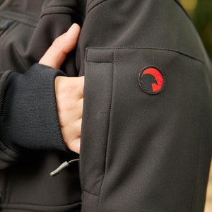 May include: Black softshell jacket with a zippered pocket on the left sleeve. The jacket has a small red logo with a stylized bird design on the sleeve.