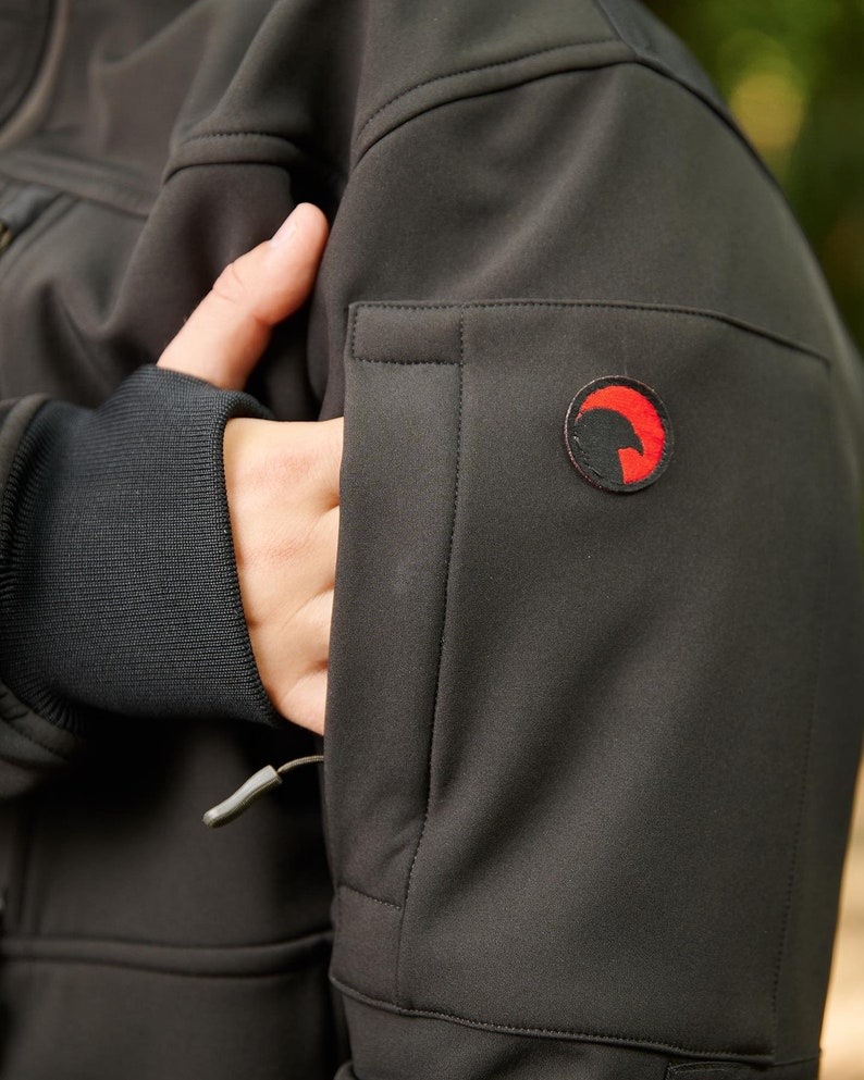 May include: Black softshell jacket with a zippered pocket on the left sleeve. The jacket has a small red logo with a stylized bird design on the sleeve.