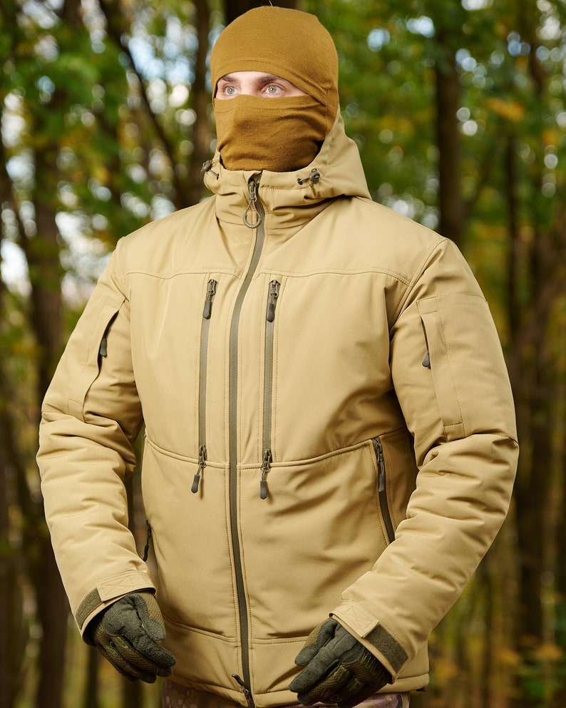 May include: A tan jacket with a zipper closure and multiple pockets. The jacket has a hood and is worn over a brown balaclava.
