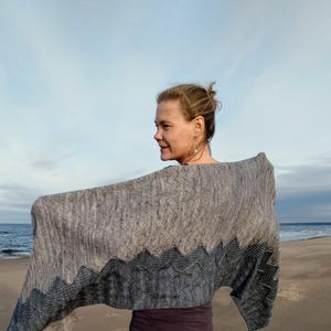May include: A woman wearing a gray and blue knitted shawl with a geometric pattern. The shawl is draped over her shoulders and she is standing on a beach.