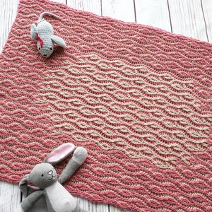 Peut inclure: Une couverture pour bébé en crochet rose et beige avec un motif ondulé. Deux jouets en peluche, une souris grise et un lapin gris, sont placés sur la couverture.