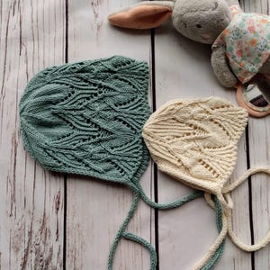 May include: Two knitted baby bonnets with ties. One bonnet is light green with a lacy pattern, the other is cream with a similar pattern.