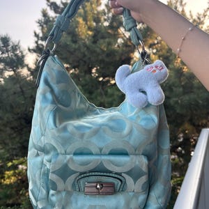 Puede incluir: Un bolso hobo de color azul claro con un patrón circular repetitivo, una sola asa trenzada y un bolsillo frontal con solapa y cierre giratorio plateado. Un pequeño dije de gato de color azul claro y textura suave cuelga del lateral.