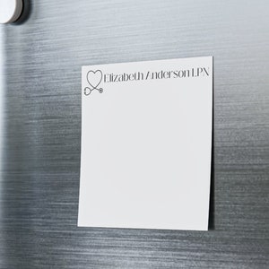 以下が含まれることがあります： 黒いハートと聴診器のデザインと「エリザベス・アンダーソン LPN」というテキストが書かれた白いメモ帳が、青い磁石でステンレス製の冷蔵庫に取り付けられています。