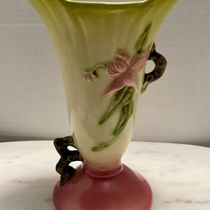 May include: A vintage ceramic vase with a green and yellow body, pink floral design, and green handles. The vase has a pink base.