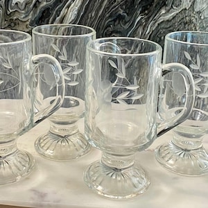May include: A set of four clear glass mugs with a pedestal base and a ribbed foot. Each mug features a delicate etched floral design with leaves and stems on the side. The mugs have a sturdy handle and a smooth, transparent finish throughout.