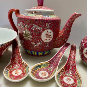 May include: A red and white porcelain tea set with a teapot, spoons, and a bowl. The teapot and spoons have a floral and swirl design. The teapot has a red handle and spout. The spoons have a white interior.