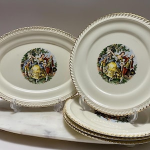 May include: A set of cream-colored ceramic plates and an oval platter featuring a central illustration of figures in period clothing. Each piece has a scalloped, gold-trimmed edge. The design depicts a group in a garden setting.