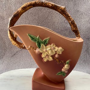 May include: A peach-colored ceramic vase with a branch-like handle. The vase features a floral design with cream-colored flowers and green leaves. The base is a darker shade of peach.