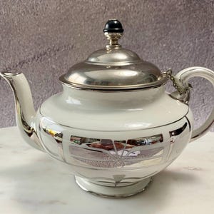 May include: A white porcelain teapot with a silver lid and silver trim. The teapot has a geometric design with a silver floral pattern.