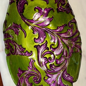 May include: A green glass bowl with a purple and white floral design. The bowl has a wavy edge and is perfect for serving snacks or holding decorative items.