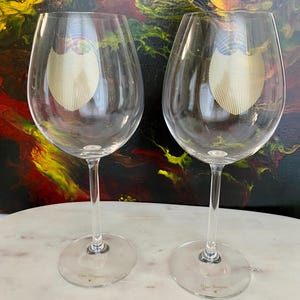 May include: Two clear wine glasses with long stems and round bases. Each glass has a gold-colored emblem on the bowl and the words "Dom Pérignon" printed on the base. The glasses are on a white surface with a colorful abstract background.