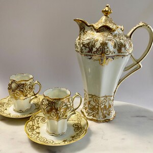May include: A white porcelain coffee pot with a matching lid and two small cups with saucers. Each piece features intricate gold filigree patterns, scalloped edges, and a glossy finish. The tall coffee pot has a curved handle and a pointed spout.