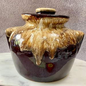 May include: A vintage ceramic bean pot with a lid. The pot is a deep brown color with a dripped glaze effect in shades of tan and brown. The pot has two side handles and a small knob on the lid.