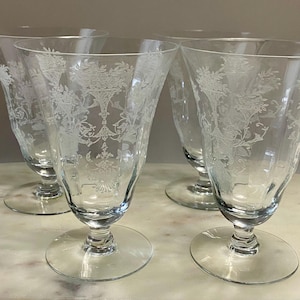 May include: Four clear, stemmed water goblets with etched floral designs. The glasses have a flared shape and sit on a round base. The etched design includes flowers and foliage. The goblets are arranged on a marble-like surface.
