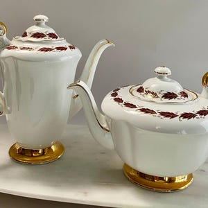 May include: Two-piece white porcelain tea set with gold accents. The set includes a teapot and a tall, slender coffee pot. Both pieces feature a gold base, handle, and rim details, and are decorated with a maroon leaf pattern around the lid.