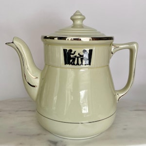 May include: Cream-colored ceramic teapot with a lid and handle, featuring a black and white silhouette design of two people at a table. The teapot has silver trim around the rim and spout.