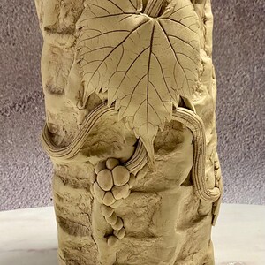 May include: A beige ceramic vase with a textured, brick-like surface. The vase features a detailed relief of a grape leaf, vines, and a cluster of grapes. The design is in a natural, earthy tone, suitable for home decor.