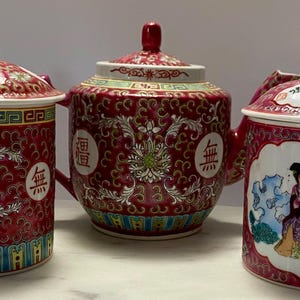 May include: Three red ceramic tea sets with lids. The sets have a floral pattern and Chinese characters. One mug has a handle and a painted scene of people. The sets are on a white surface.