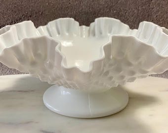May include: A white, ruffled-edge bowl with a pedestal base. The bowl has a textured, hobnail-like pattern on the exterior and a smooth interior. The ruffled edge adds a decorative touch to this elegant serving piece.
