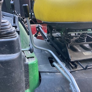 May include: A close-up of a green tractor seat with a silver lever. The lever is attached to the seat and has a black knob. The seat is yellow and has a black base.