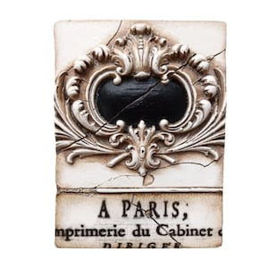 Könnte beinhalten: Rechteckige, cremefarbene Dekorplatte mit schwarzem, ovalem Mittelteil und cremefarbenen Schnörkeln. Die Platte ist gerissen und trägt unten den Text "A PARIS; imprimerie du Cabinet".