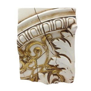 Könnte beinhalten: Fragment eines dekorativen architektonischen Elements. Das Stück hat eine cremefarbene Basis mit goldenen Akzenten und aufwendigen floralen und scrollartigen Designs. Die Oberfläche ist rissig und wirkt dadurch gealtert.