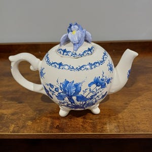 Könnte beinhalten: Eine weiße Keramik-Teekanne mit blauem Blumenmuster und einer skulptierten Irisblüte auf dem Deckel. Die Teekanne hat einen geschwungenen Griff, einen Ausguss und steht auf drei kleinen Füßen. Der Körper ist mit detaillierten blauen Irisblüten verziert.