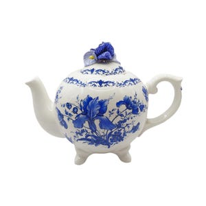Könnte beinhalten: Weiße Keramik-Teekanne mit blauen Blumenmustern, darunter Iris und andere Blumen. Die Teekanne hat einen gebogenen Ausguss, einen Henkel und drei kleine Füße. Eine blaue Blume verziert den Deckel.