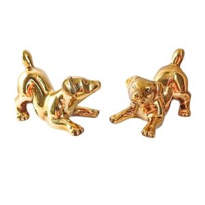 Peut inclure: Deux figurines de chiens dorés dans des poses ludiques. Les chiens sont sculptés avec des détails et une finition brillante. Les figurines sont posées sur un fond blanc, soulignant leur éclat métallique.