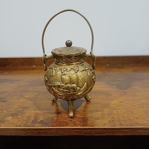 May include: A small, round, brass-colored metal pot with a lid, a swing handle, and three legs. The exterior features a textured surface with a relief design of a sailing ship and foliage. The lid has a small central knob.