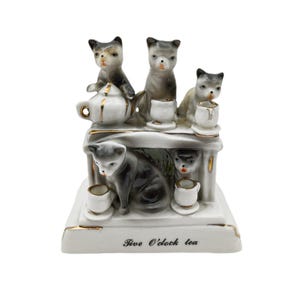 May include: A ceramic figurine featuring five gray and white cats around a tea set. The cats are posed with teacups and a teapot. The base of the figurine has the text "Five O'clock tea" in black script, with gold trim.
