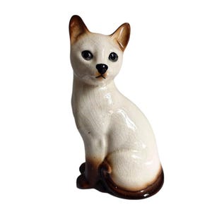 May include: A ceramic cat figurine in a seated position with a cream-colored body and dark brown accents on the ears, face, and tail. The surface features a visible crackle glaze finish, with dark eyes and a small black nose on the feline face.