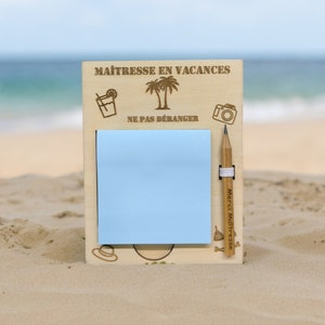 Peut inclure: Un panneau en bois avec le texte "Maîtresse en vacances" et "Ne pas déranger" avec un post-it bleu et un crayon attaché. Le panneau est sur une plage de sable.