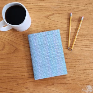 May include: A blue notebook with a woven pattern sits on a wooden table with a cup of coffee and two yellow pencils.