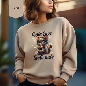 Può includere: Una felpa color sabbia con un'illustrazione di un gatto nero e arancione e il testo "Gotta Love Torti-tude".