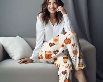 Puede incluir: Pantalón de pijama blanco con un estampado de temática otoñal que incluye calabazas, tazas de café, corazones a cuadros, girasoles y hojas. El texto "Fall in the air" está impreso en el pantalón.