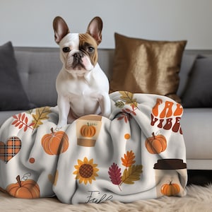 May include: A white and brown French bulldog dog sitting on a white throw blanket with a fall-themed design. The blanket features pumpkins, leaves, sunflowers, coffee cups, and the text "Fall Vibes".