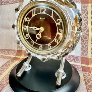 May include: A vintage crystal desk clock with a black base and silver supports. The clock face is brown with gold numerals and hands, and the word "Majak" is printed on the face. The clock is marked "Made in USSR".